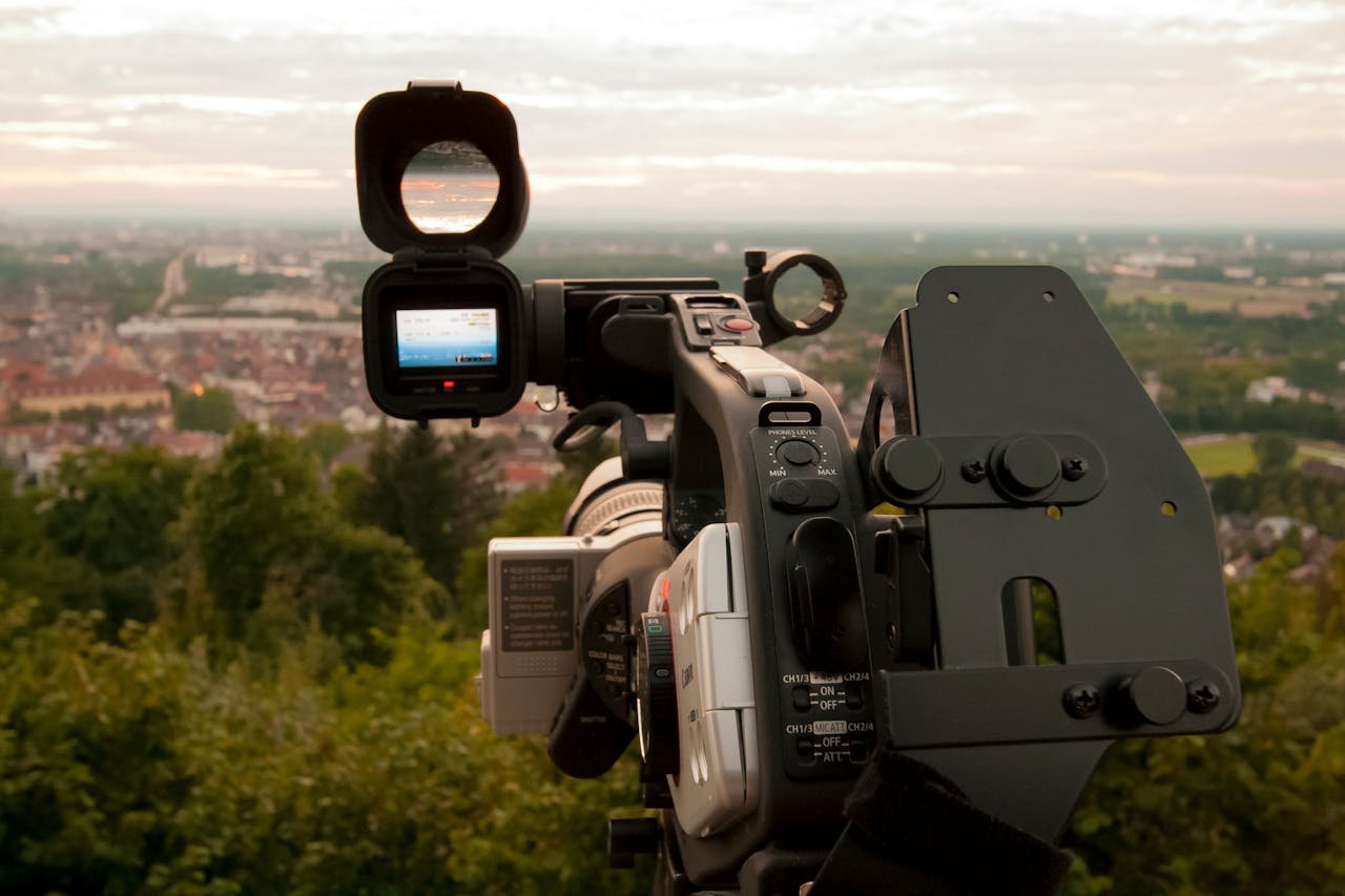 our-services-03 Close-up of a professional video camera set up outdoors with a city skyline in the background.